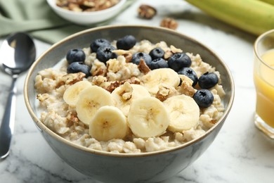 Photo of Tasty oatmeal with banana, blueberries, walnuts and milk served in bowl on white marble table