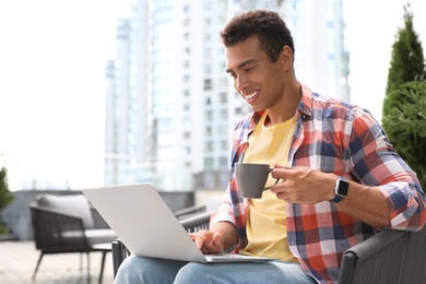 Portrait of handsome young African-American man with laptop and cup of drink in outdoor cafe