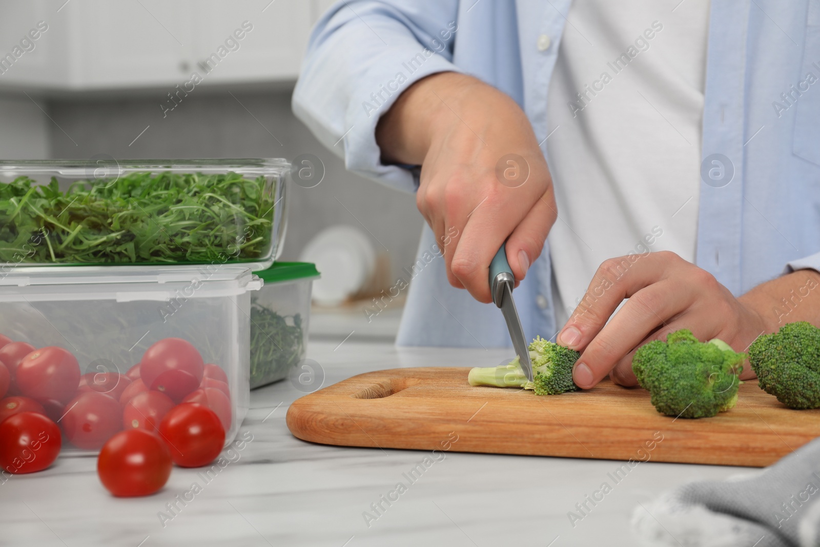 Photo of Man cutting fresh broccoli with knife near containers at white marble table in kitchen, closeup. Food storage