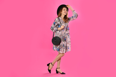 Photo of Young woman wearing floral print dress with stylish handbag and hat on pink background