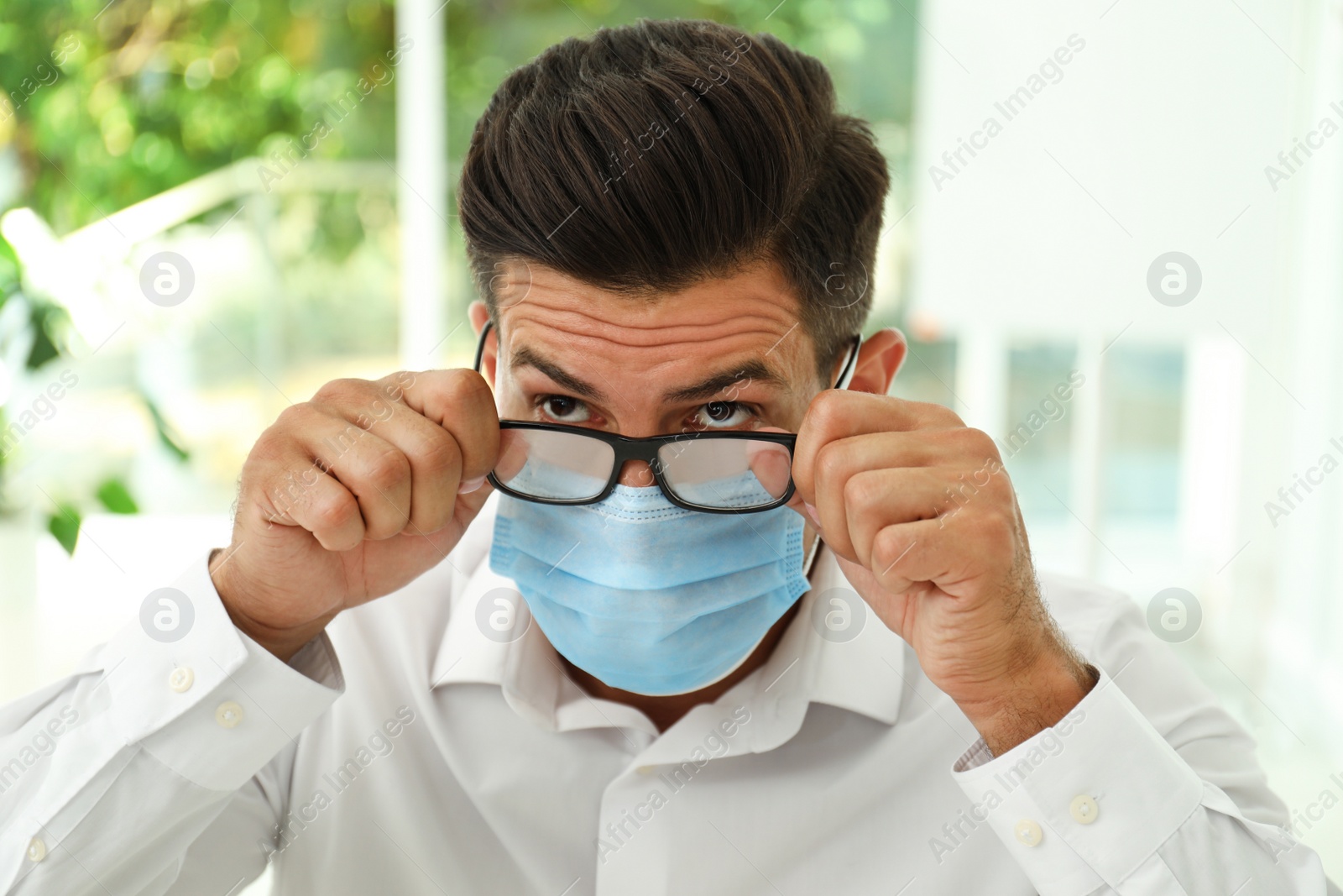 Photo of Man wiping foggy glasses caused by wearing medical mask indoors, closeup