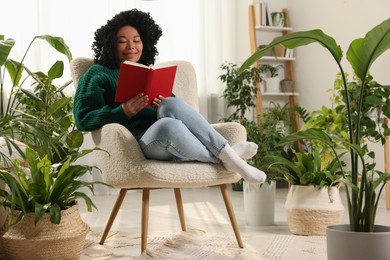 Photo of Relaxing atmosphere. Happy woman reading book on armchair surrounded by beautiful houseplants in room
