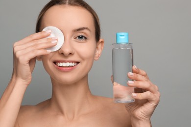 Smiling woman removing makeup with cotton pad and holding bottle on grey background, closeup