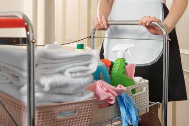 Photo of Young chambermaid with cart and cleaning products in hotel, closeup