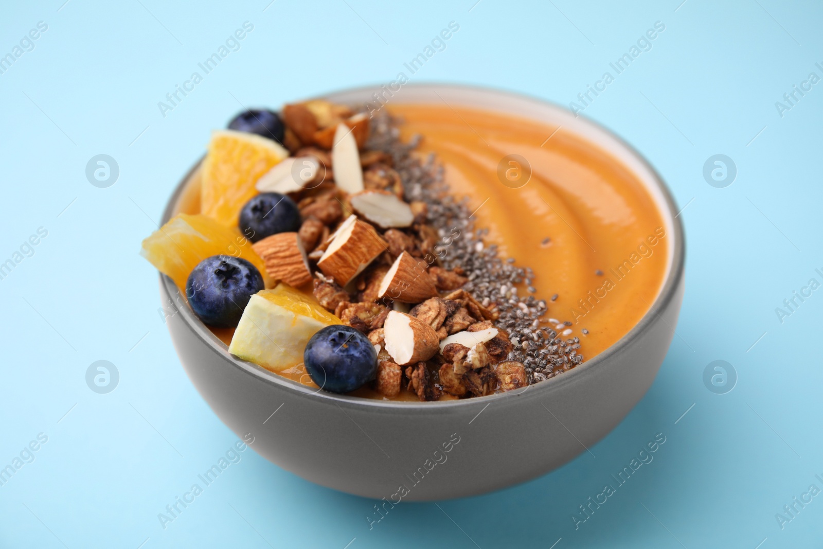 Photo of Bowl of delicious fruit smoothie with fresh orange slices, blueberries and granola on light blue background, closeup