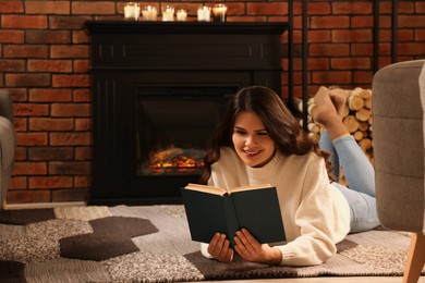 Photo of Young woman reading book on floor near fireplace at home