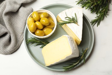 Photo of Plate with pieces of tasty camembert cheese, olives and rosemary on white table, top view