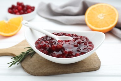 Photo of Fresh cranberry sauce in bowl, spoon and rosemary on white wooden table, closeup