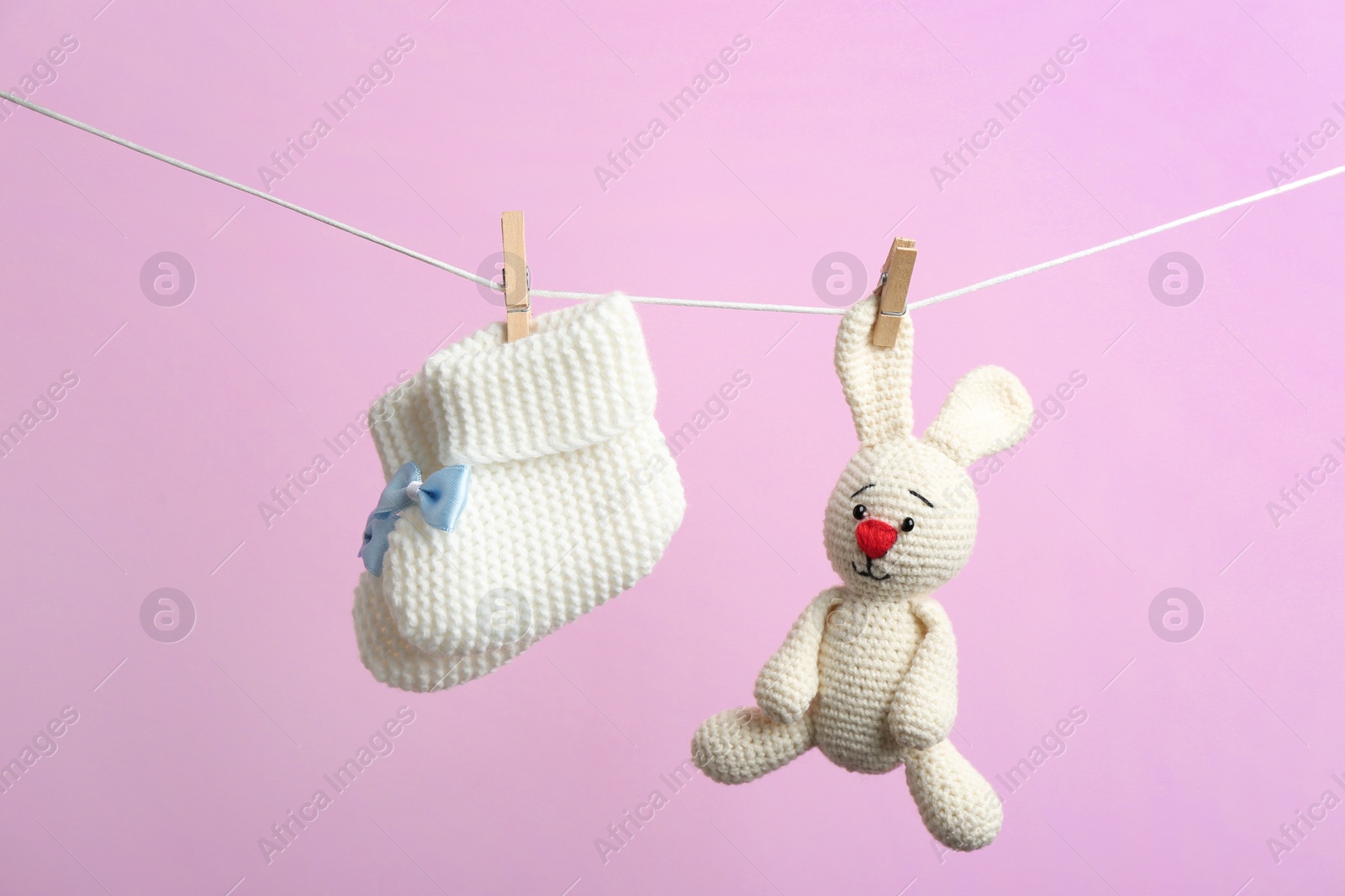 Photo of Knitted booties and toy bunny hanging on washing line against color background. Baby accessories