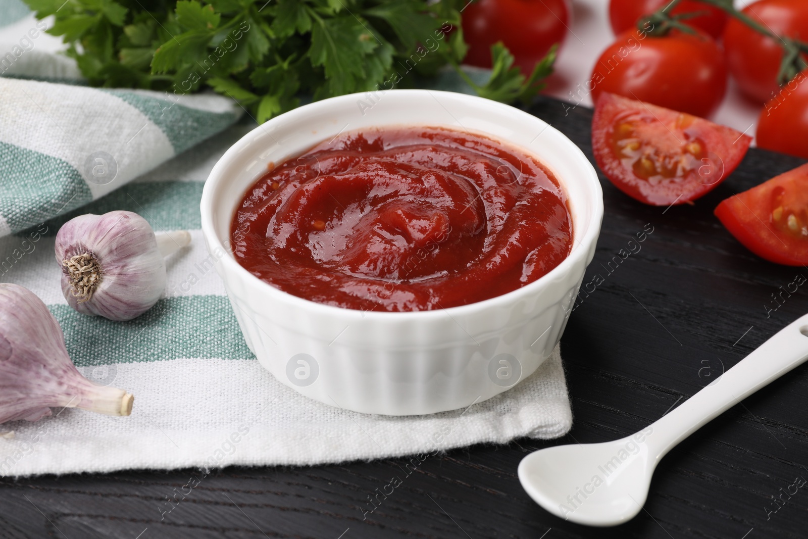 Photo of Organic ketchup in bowl, fresh tomatoes and garlic on black table, closeup. Tomato sauce