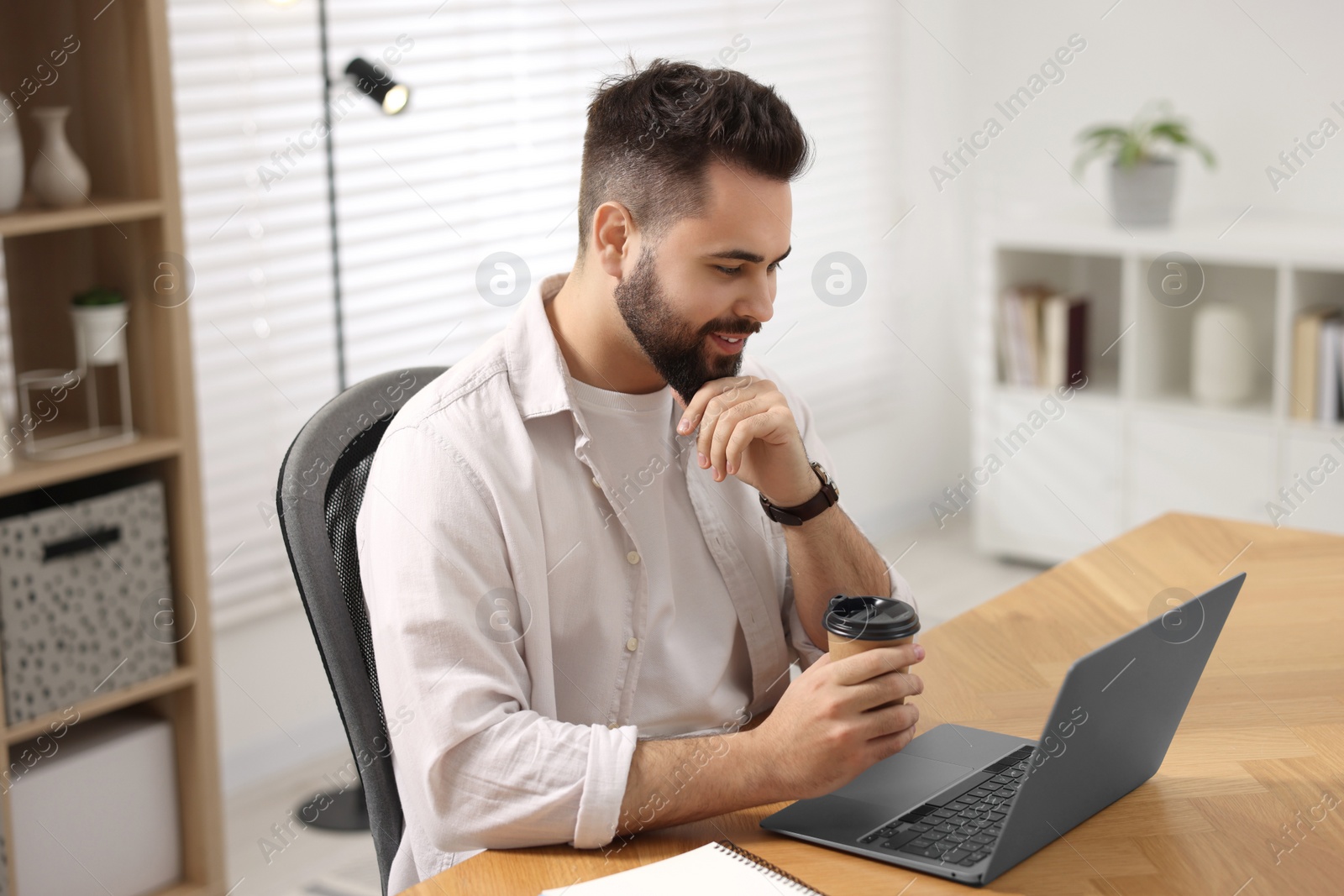 Photo of Young man with cup of coffee watching webinar at table in room