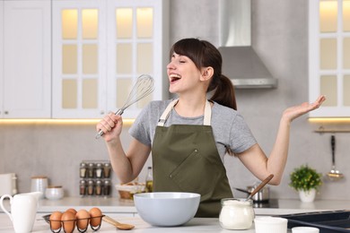 Photo of Happy young housewife with whisk having fun while cooking at white table in kitchen