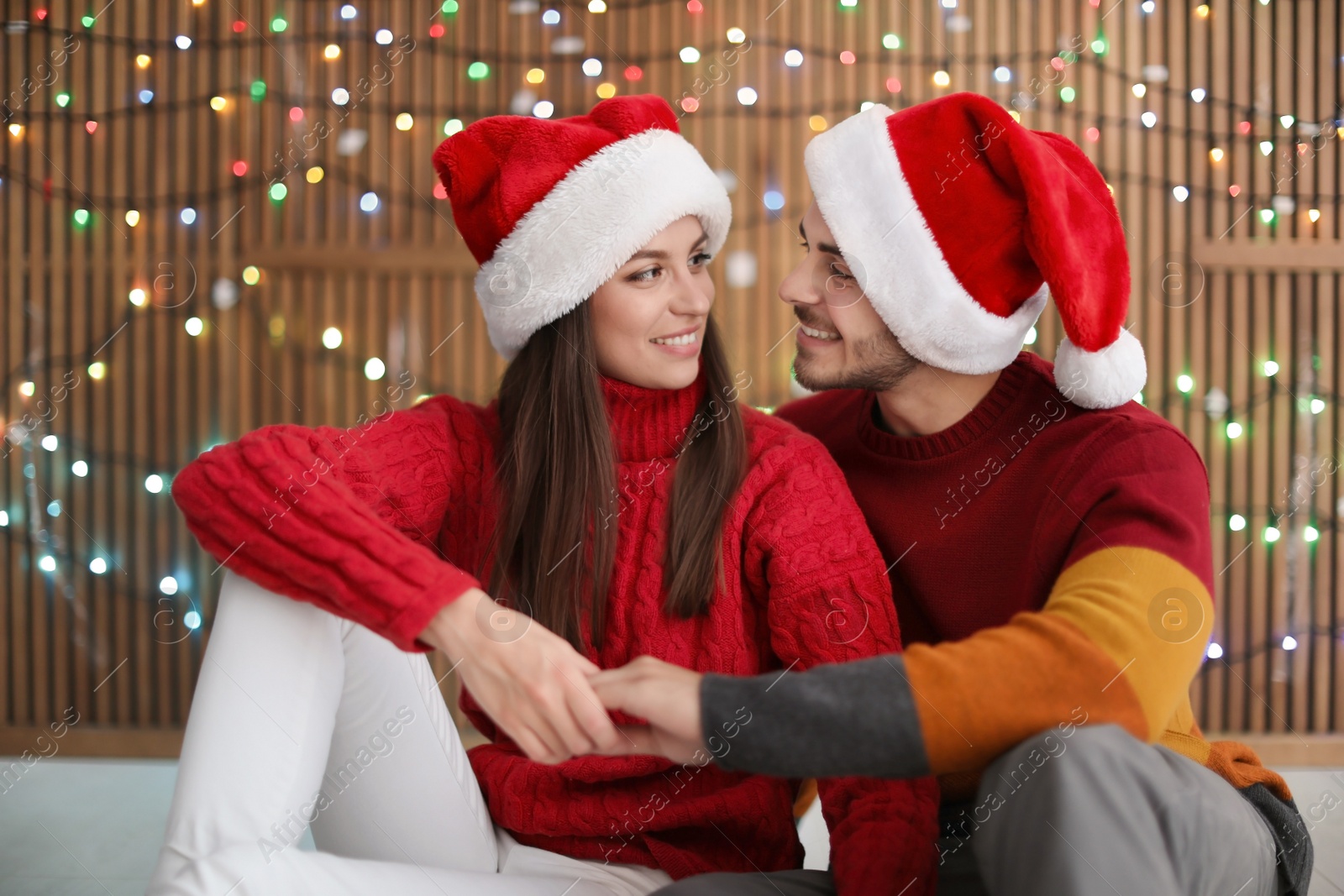 Photo of Young couple in Santa hats on blurred lights background. Christmas celebration
