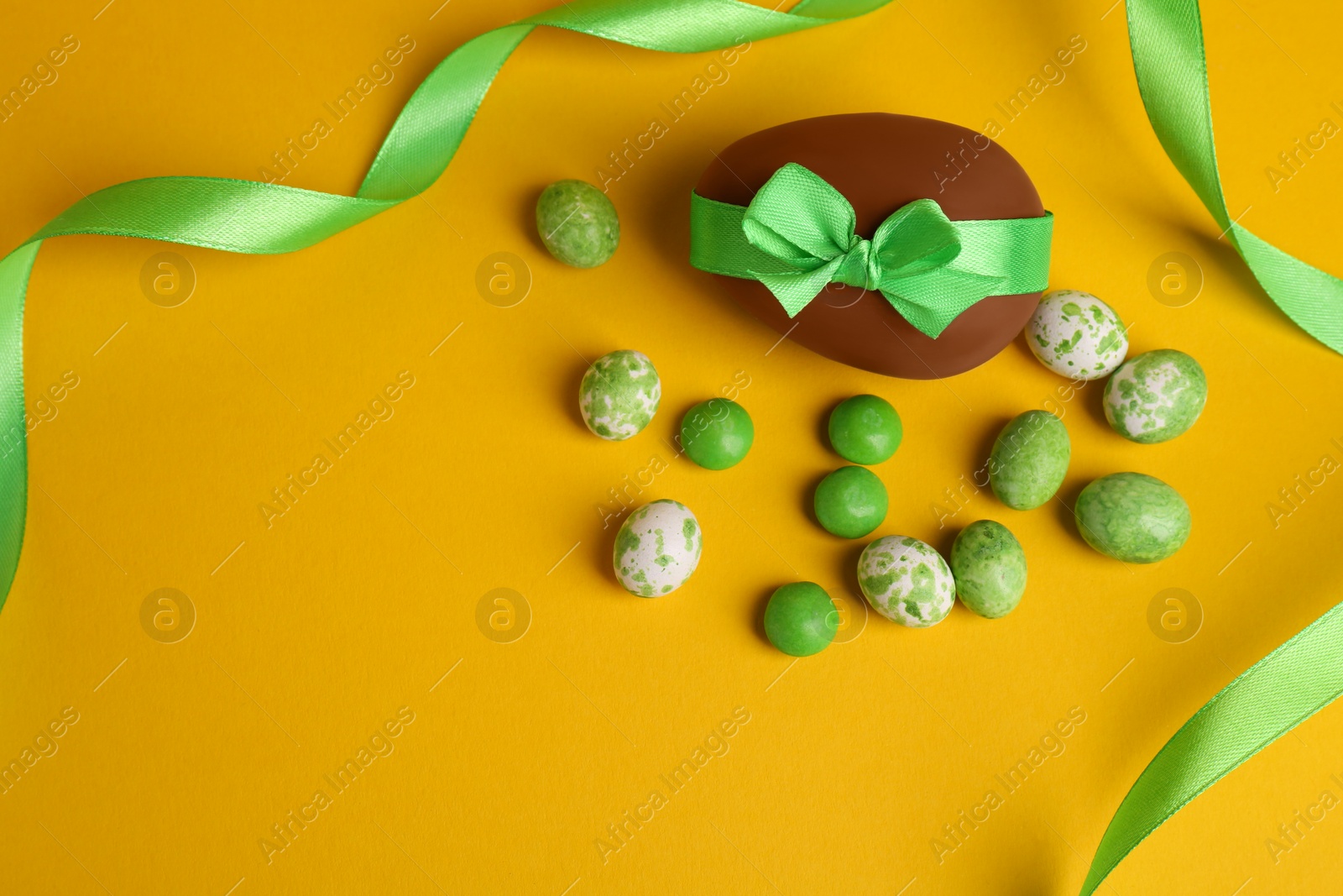 Photo of Tasty chocolate egg with green ribbon and candies on yellow background, flat lay. Space for text