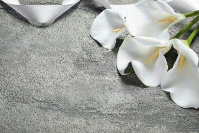 Photo of Beautiful calla lily flowers and white ribbon on grey table, space for text. Funeral symbols