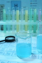 Beaker with liquid, test tubes and magnifying glass on periodic table of chemical elements. Light blue tone effect