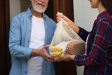 Photo of Helping neighbours. Young woman with net bag of products visiting senior man outdoors, closeup