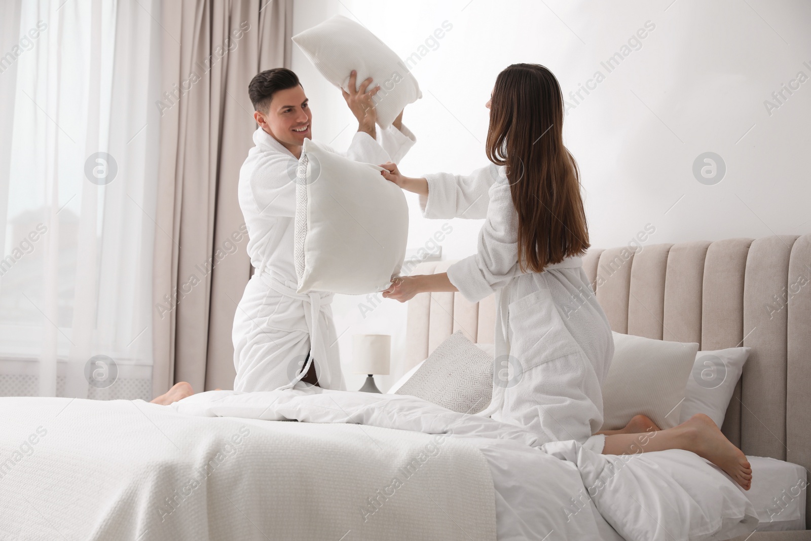 Photo of Happy couple in bathrobes having pillow fight on bed at home