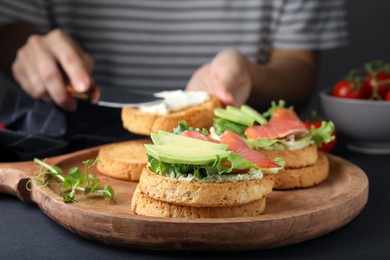 Photo of Woman making tasty rusks with salmon, cream cheese and avocado at black table, closeup