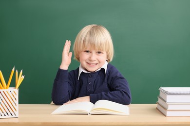 Happy little school child raising hand while sitting at desk with books near chalkboard