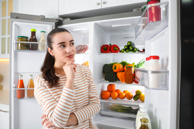 Photo of Thoughtful young woman near open refrigerator indoors