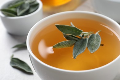 Photo of Cup of sage tea with green leaves on white table, closeup