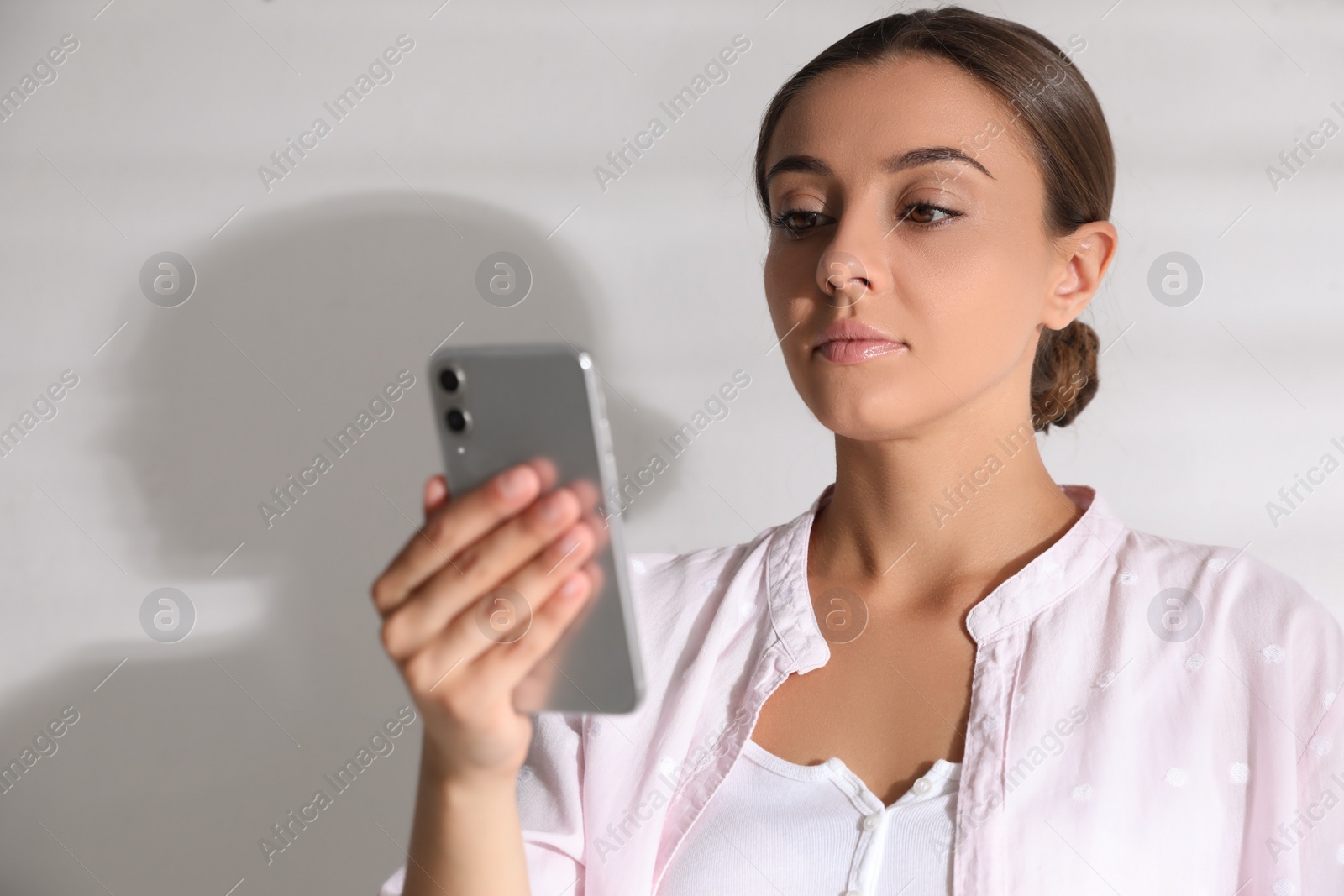 Photo of Young woman unlocking smartphone with facial scanner near white wall. Biometric verification