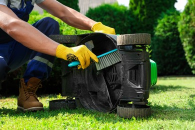 Young man cleaning lawn mower with brush in garden, closeup