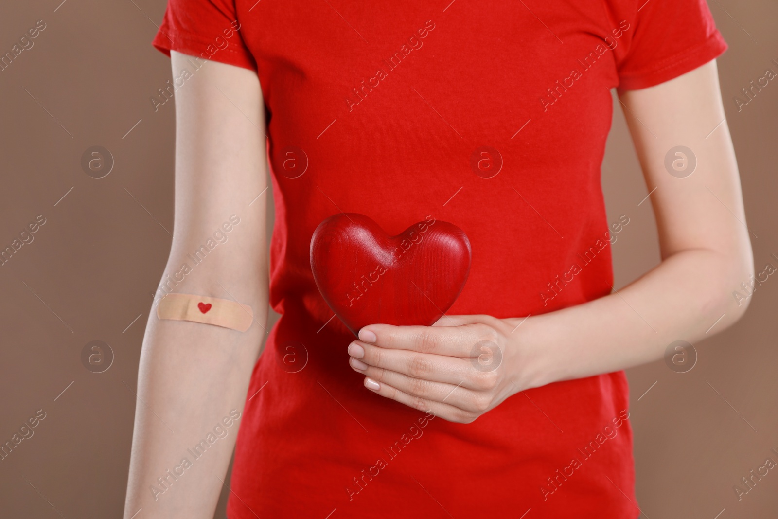 Photo of Blood donation concept. Woman with adhesive plaster on arm holding red heart against brown background, closeup