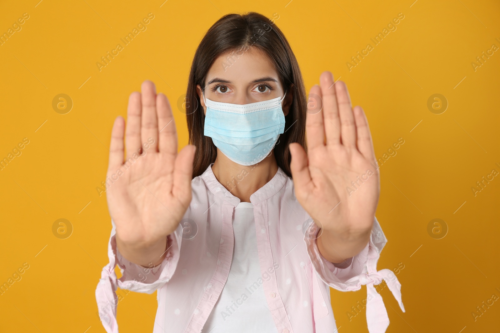 Photo of Woman in protective mask showing stop gesture on yellow background. Prevent spreading of coronavirus