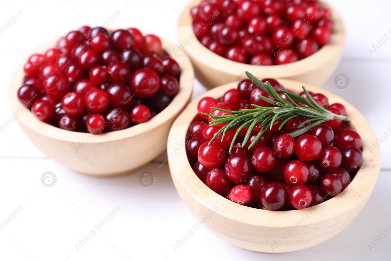 Photo of Fresh ripe cranberries in bowls and rosemary on white wooden table, closeup