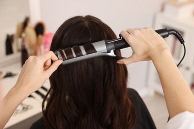Photo of Hairdresser working with client using curling hair iron in salon, closeup