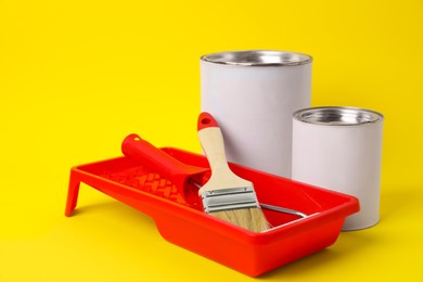 Cans of orange paint, brush, roller and container on yellow background