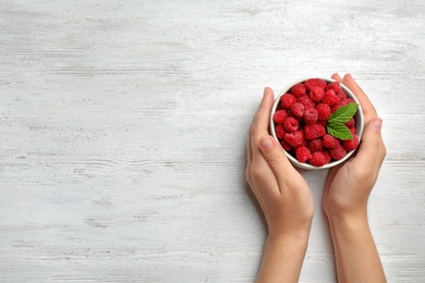 Photo of Woman holding bowl with delicious ripe raspberries at white wooden table, top view. Space for text