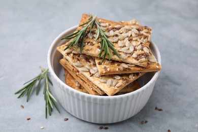 Photo of Cereal crackers with flax, sunflower, sesame seeds in bowl and rosemary on grey table, closeup