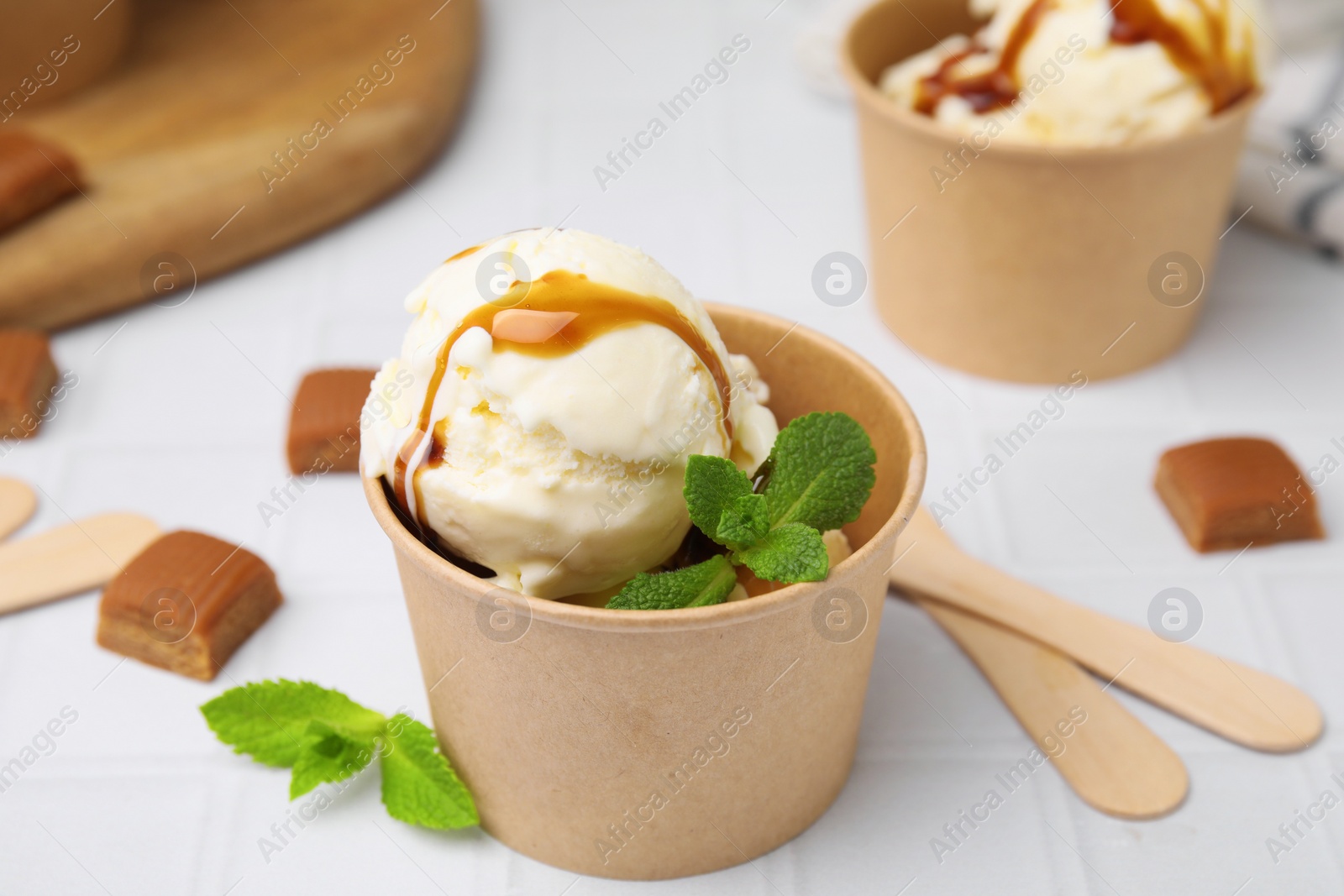 Photo of Scoops of ice cream with caramel sauce, mint leaves and candies on white tiled table, closeup