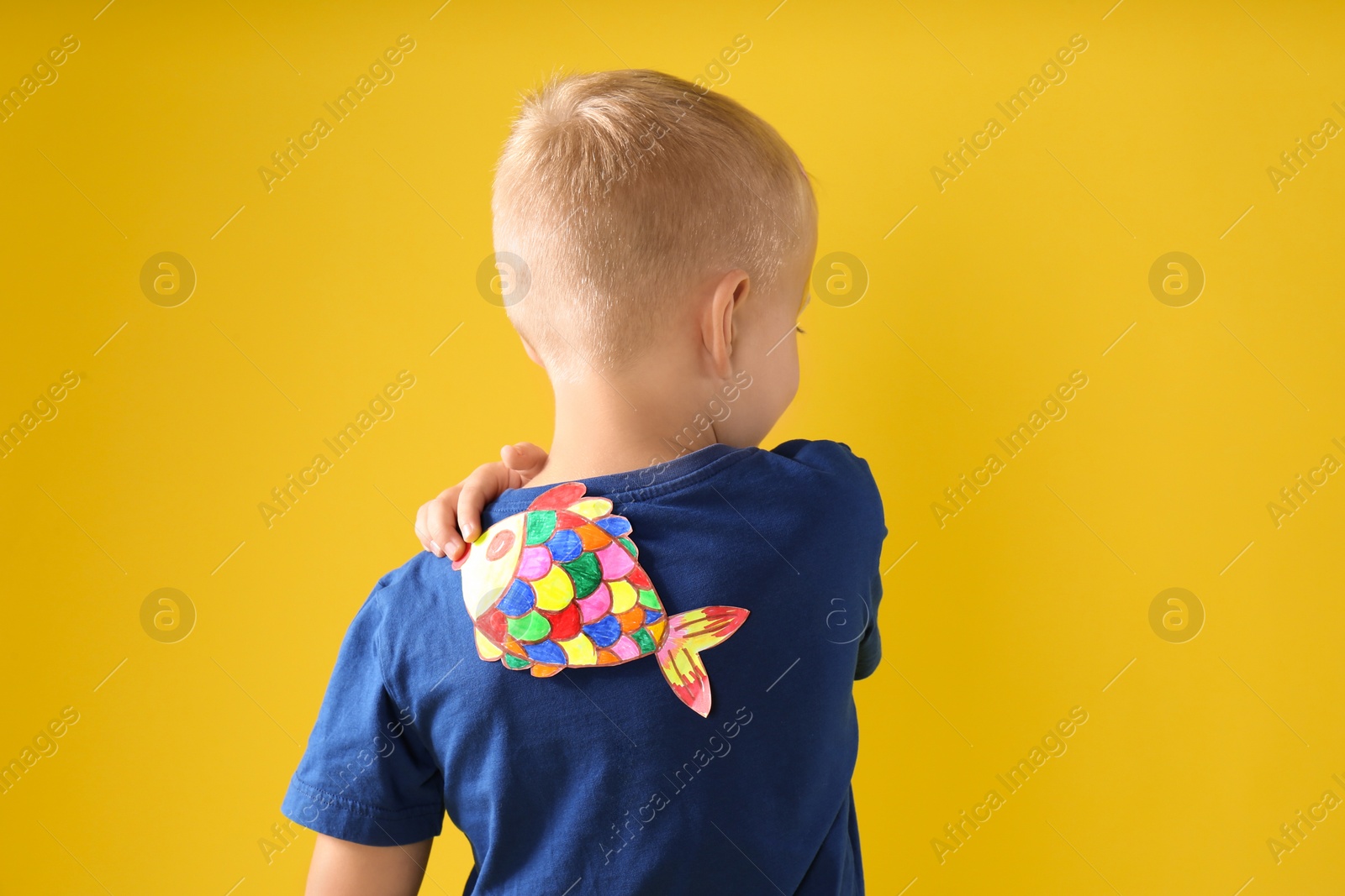 Photo of Little boy with paper fish on back against yellow background. April fool's day