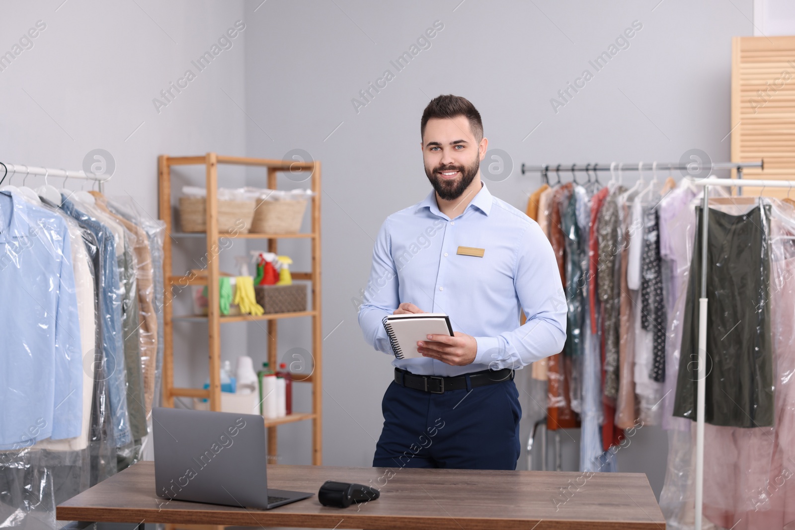 Photo of Dry-cleaning service. Happy worker with notebook in workplace indoors