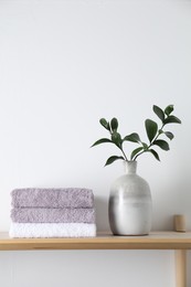 Photo of Stacked terry towels and green branches in vase on wooden shelf near white wall