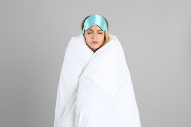 Photo of Young woman in sleeping mask wrapped with soft blanket on grey background