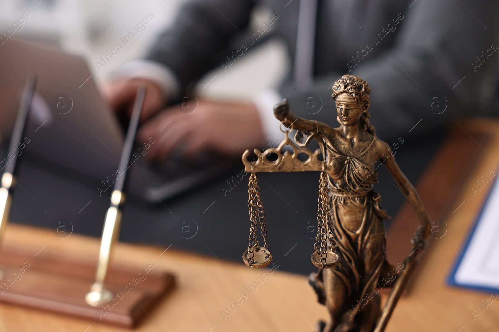 Photo of Notary working with laptop at wooden table in office, focus on statue of Lady Justice