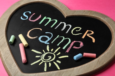 Photo of Heart shaped blackboard with text SUMMER CAMP, drawing and chalk sticks on color background