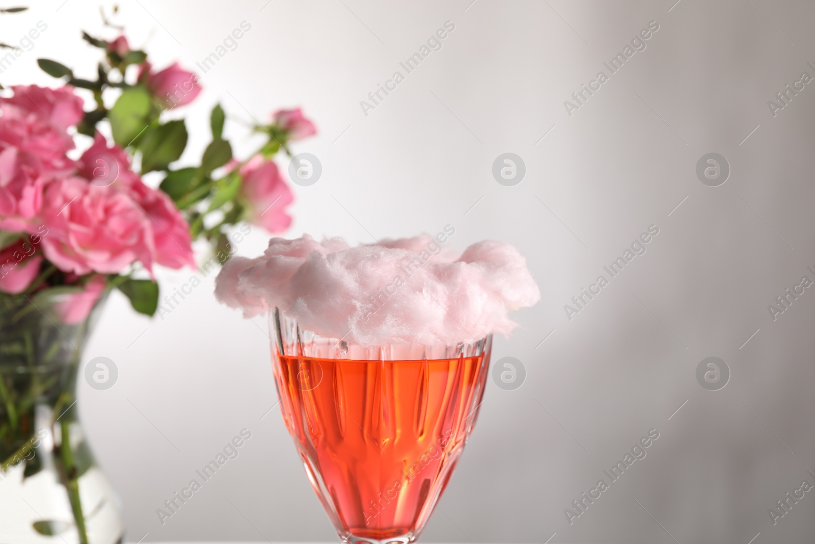 Photo of Cotton candy cocktail in glass and vase with pink roses on gray background, closeup. Space for text