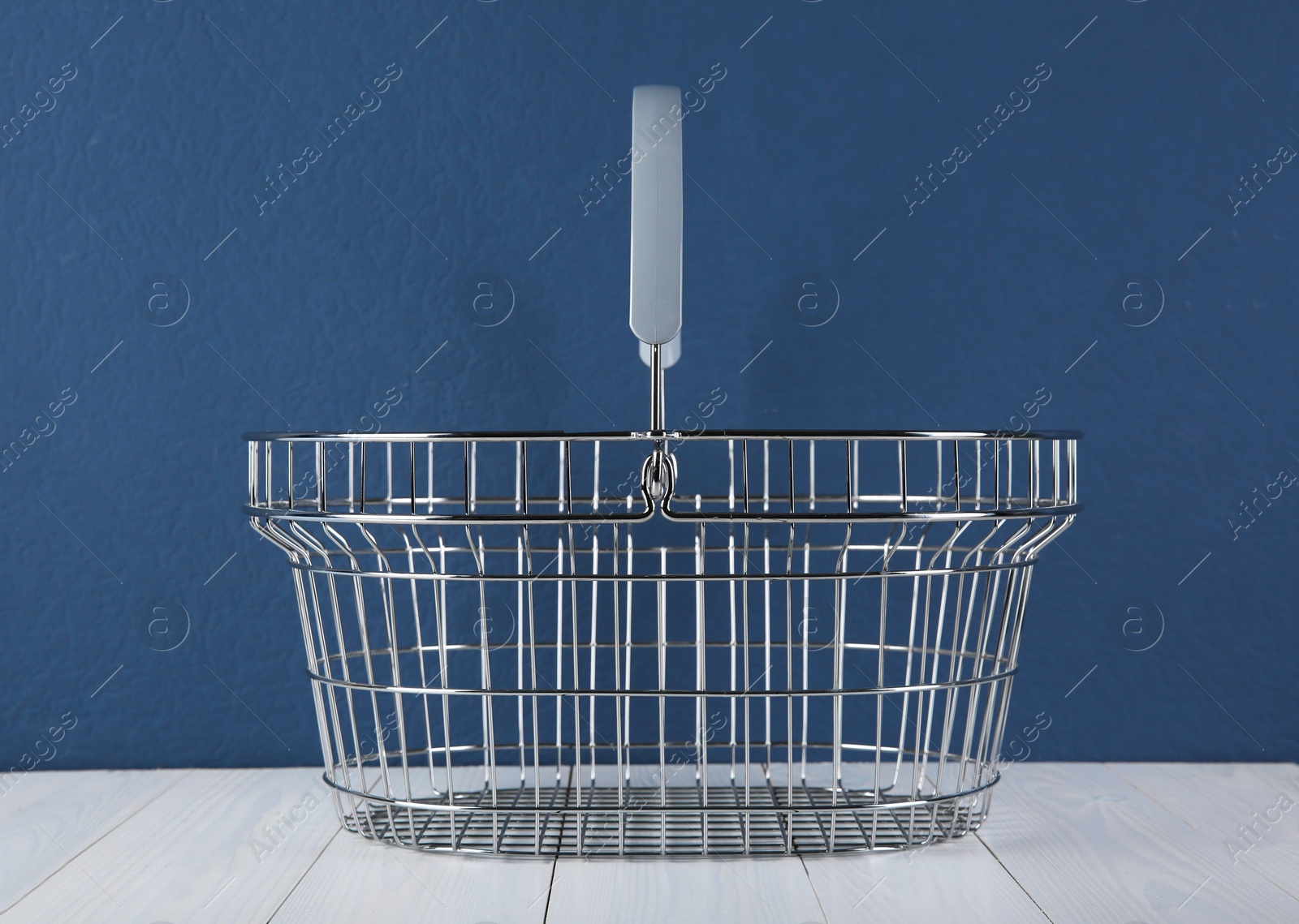 Photo of Empty metal shopping basket on white wooden table against blue background