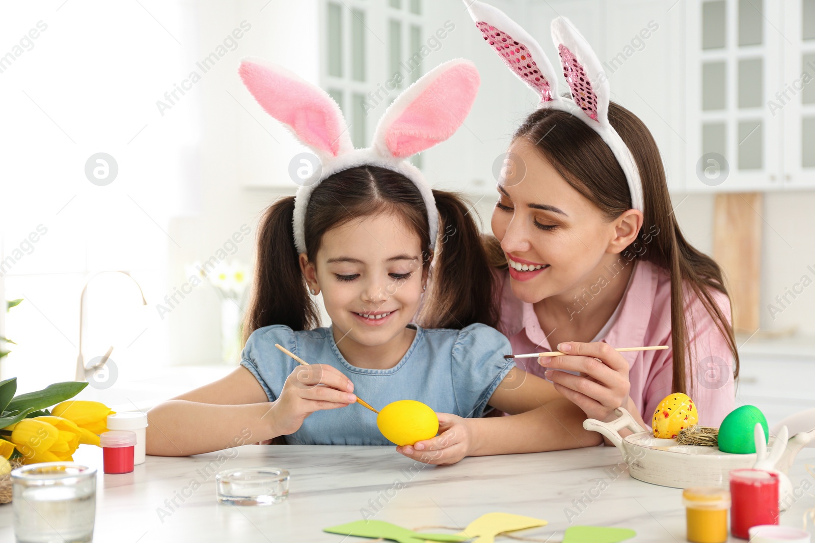 Photo of Happy mother and daughter with bunny ears headbands painting Easter egg in kitchen