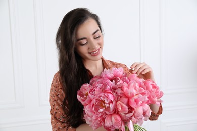 Beautiful young woman with bouquet of pink peonies near white wall