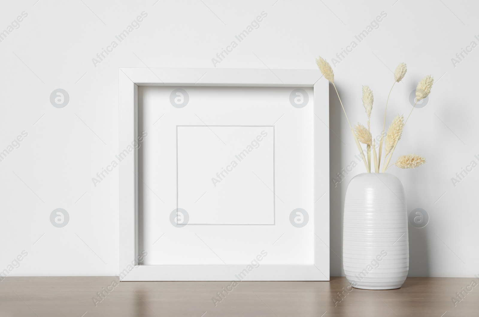 Photo of Empty photo frame and vase with dry decorative spikes on wooden table. Mockup for design