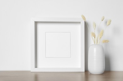 Photo of Empty photo frame and vase with dry decorative spikes on wooden table. Mockup for design