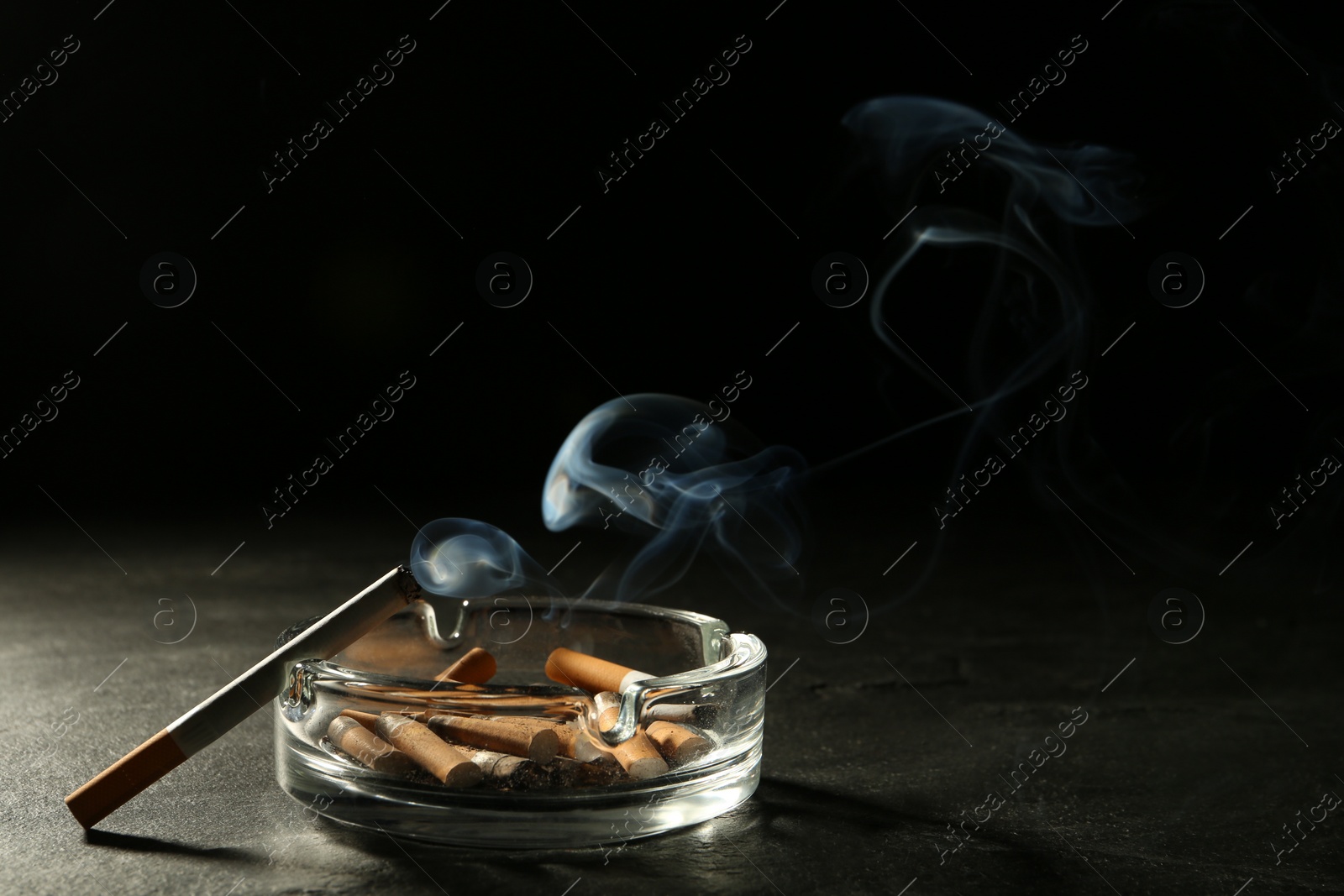 Photo of Glass ashtray with stubs and smoldering cigarette on grey table against black background. Space for text