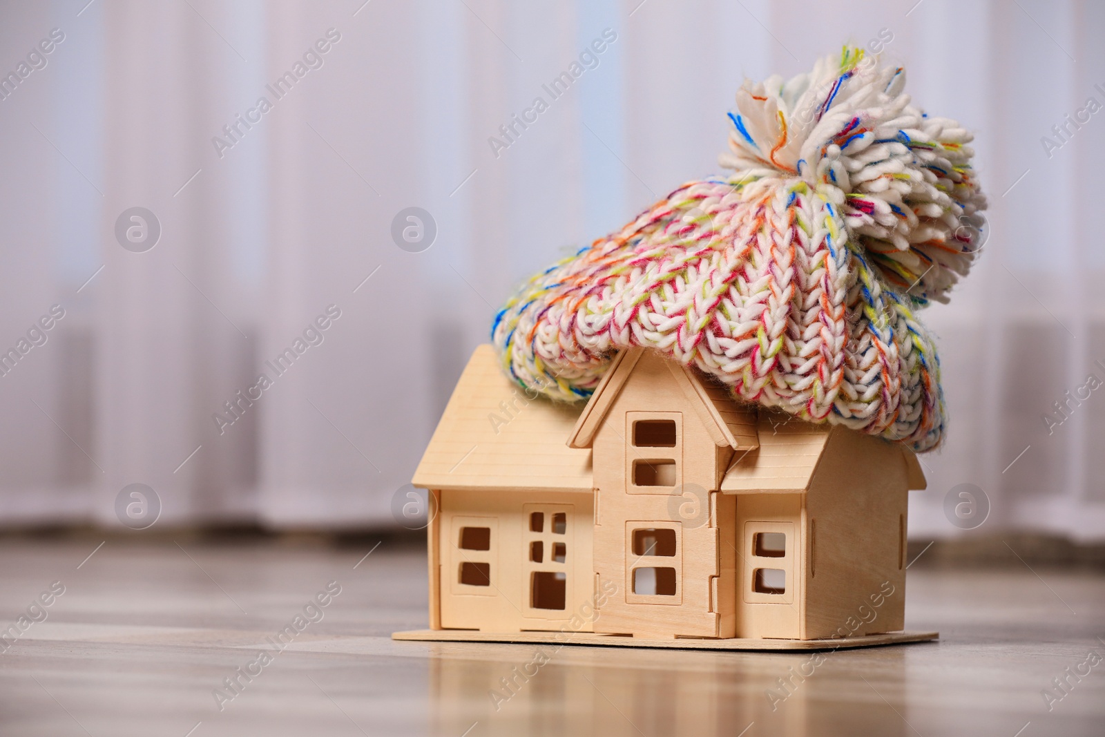 Photo of Wooden house model in hat on floor indoors, space for text. Heating efficiency